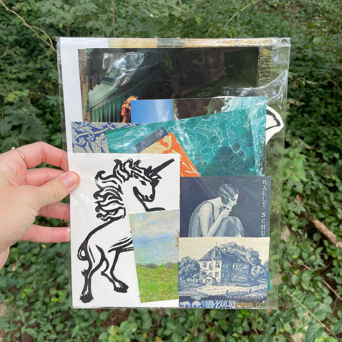 A hand holds a clear plastic sleeve containing a collage of printed art pieces, including a black line drawing of a unicorn, a blue landscape, and a crouching figure, against a background of green foliage.