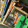 A close-up, angled view of a stack of vintage illustrated postcards or art prints fanned out on a textured green surface. The prints feature richly detailed, colorful fantasy and botanical illustrations, including a prominent green card with a black sword design and a blue-and-orange card with a winged figure.