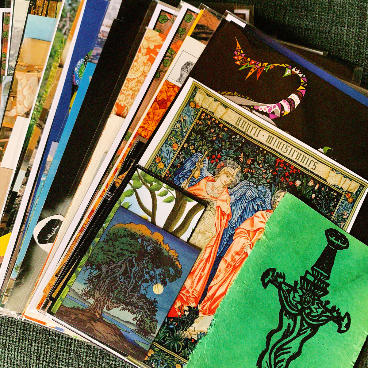 A close-up, angled view of a stack of vintage illustrated postcards or art prints fanned out on a textured green surface. The prints feature richly detailed, colorful fantasy and botanical illustrations, including a prominent green card with a black sword design and a blue-and-orange card with a winged figure.