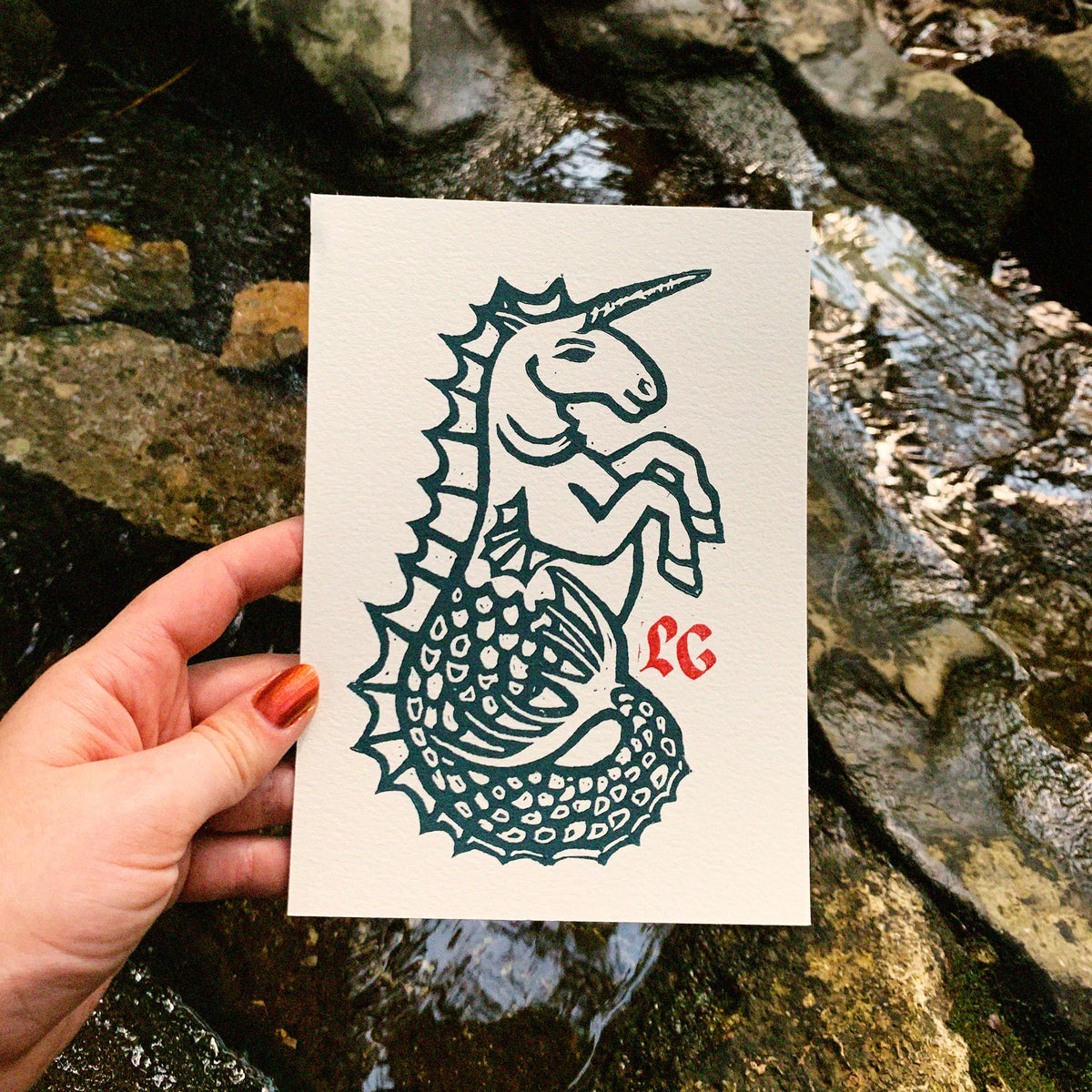 A hand holds a white paper art print featuring a dark teal linoleum print of a unicorn with a mermaid tail and a small red monogram in the corner, set against a natural rocky stream background.