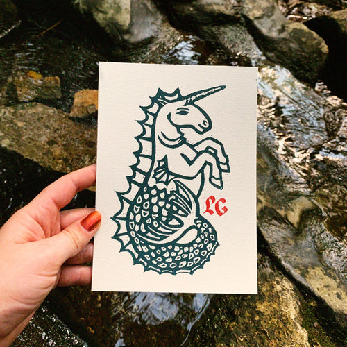 A hand holds a white paper art print featuring a dark teal linoleum print of a unicorn with a mermaid tail and a small red monogram in the corner, set against a natural rocky stream background.
