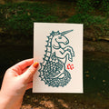 A hand with orange nail polish holds a white rectangular art print featuring a dark teal hippocampus illustration with a small red 'LG' monogram in the corner, set against an outdoor background of greenery and a Kentucky blue hole.