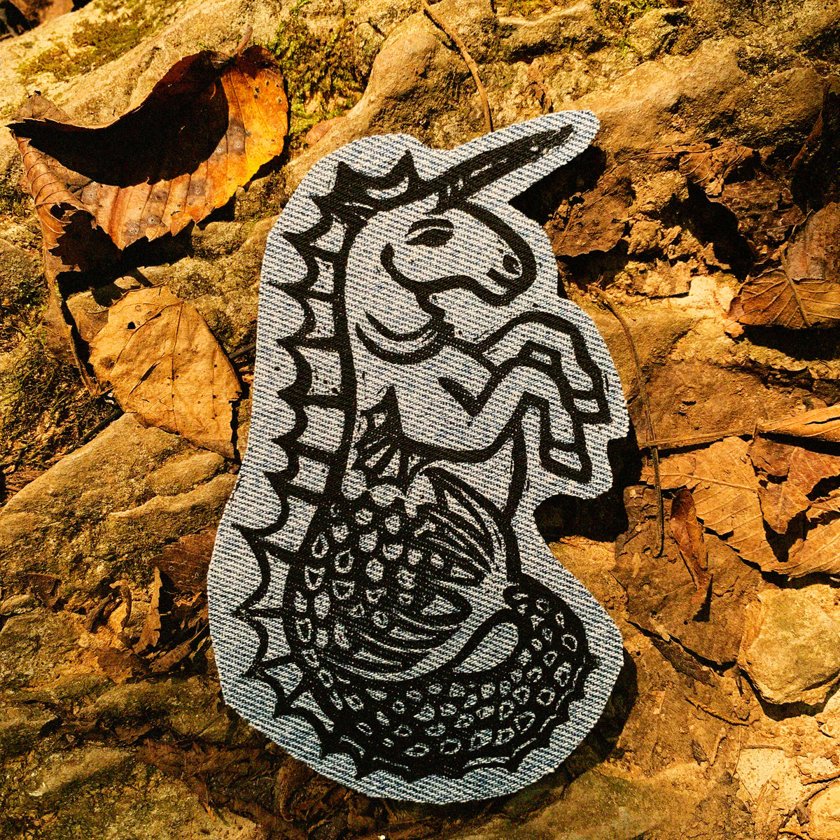 A hippocampus-shaped fabric patch featuring a black ink-style illustration of the traditional unicorn with a fish tail, placed on a background of dry brown leaves and rocks.