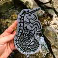 A hand holds a patch featuring a black linocut print of a hippocampus on a denim fabric patch, set against a natural rocky stream backdrop.