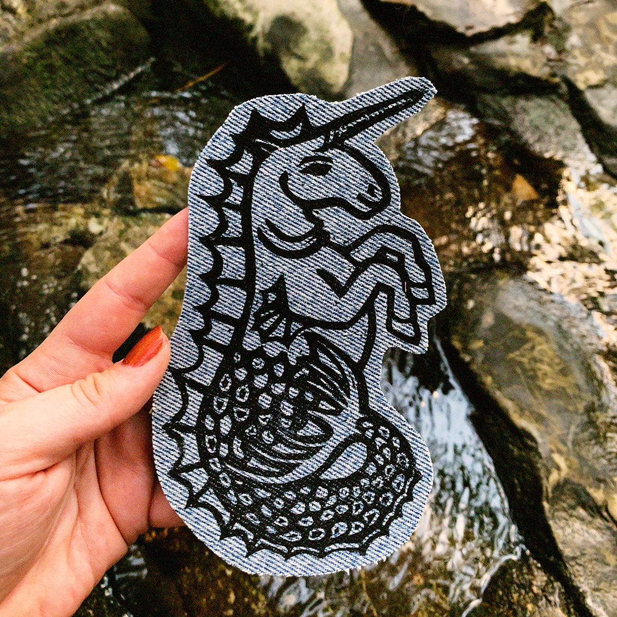 A hand holds a patch featuring a black linocut print of a hippocampus on a denim fabric patch, set against a natural rocky stream backdrop.