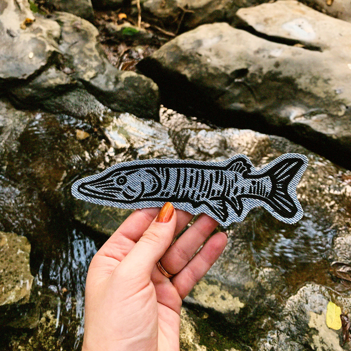 A hand holds a fish-shaped patch with a black Muskie fish design on a blue denim fabric background, set against a natural outdoor background of rocks and flowing water.