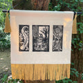 A rectangular cream canvas wall hanging with three black woodblock-style vertical print panels and gold fringe along the bottom, suspended outdoors from a black rod with two gold tassels on either side. The product is photographed against a real outdoor background with tree bark and green foliage visible.