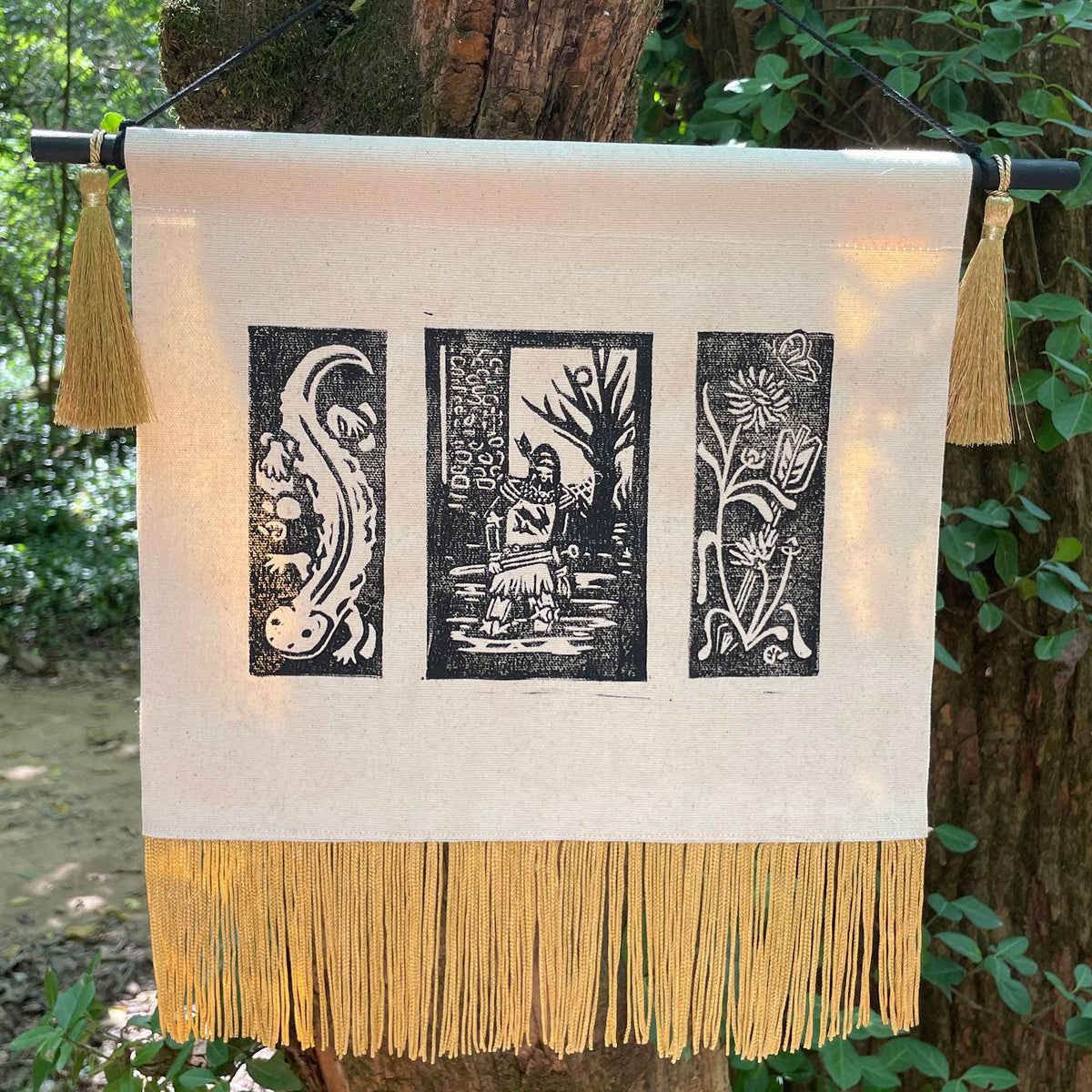 A rectangular cream canvas wall hanging with three black woodblock-style vertical print panels and gold fringe along the bottom, suspended outdoors from a black rod with two gold tassels on either side. The product is photographed against a real outdoor background with tree bark and green foliage visible.