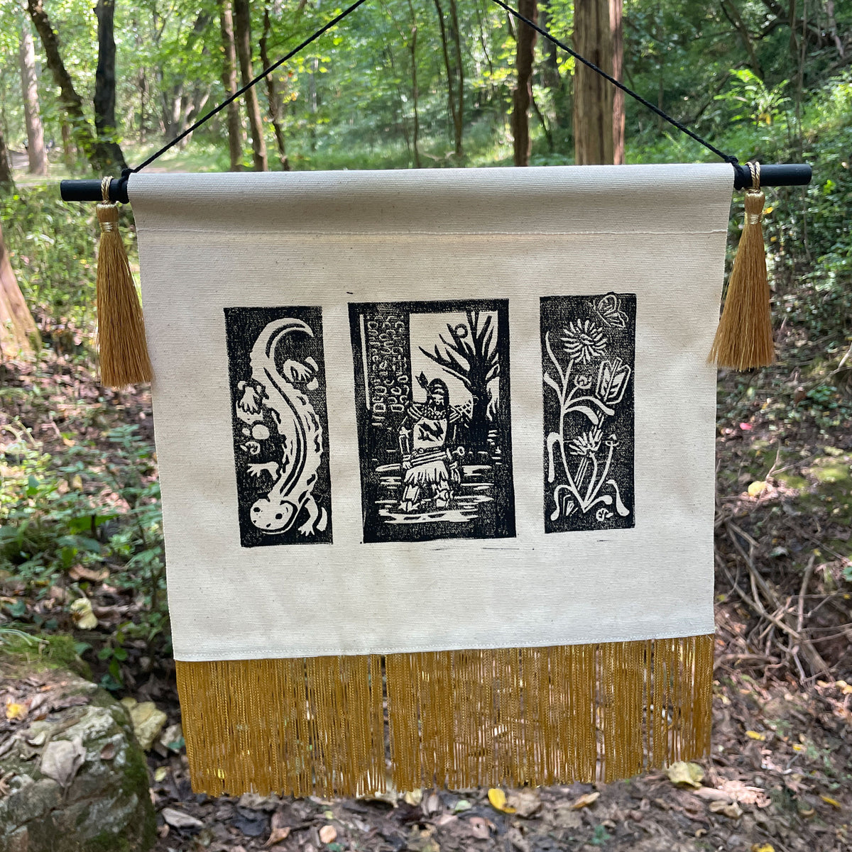 A canvas wall hanging tapestry with three black woodblock-style panels featuring a salamander, a knight under a tree, and a flower and arrow, suspended from a black rod outdoors in a forest setting. The tapestry has a cream background, a gold tassel fringe at the bottom, and gold tassels on the sides.