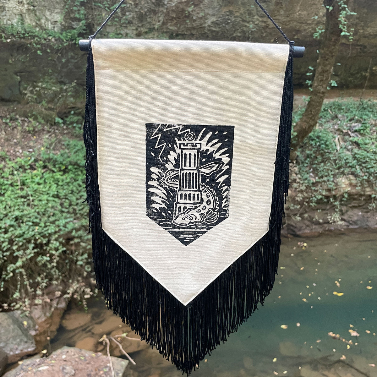 A vertical fabric banner with black fringe hangs outdoors near a stream and trees. The banner has a natural canvas center panel with a black printed crest showing a tower and a sturgeon, suspended from a black rod.