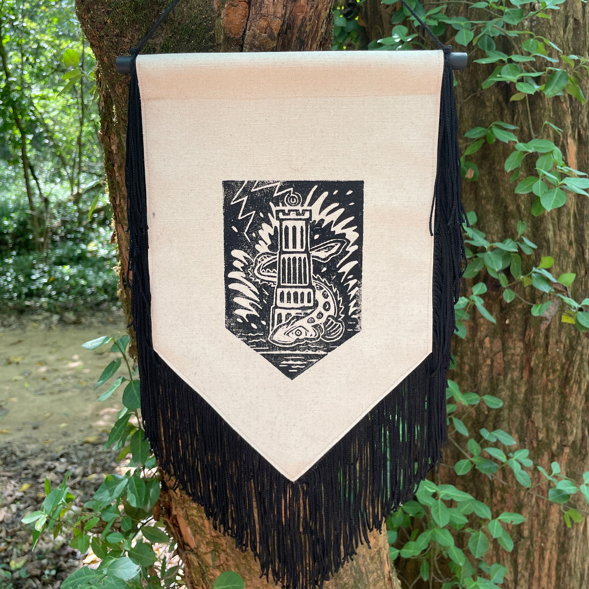 A vertical canvas banner with black fringe hangs from a tree outdoors. The banner is cream-colored with a black printed shield graphic in the center featuring a tower, lightning, and a sturgeon.