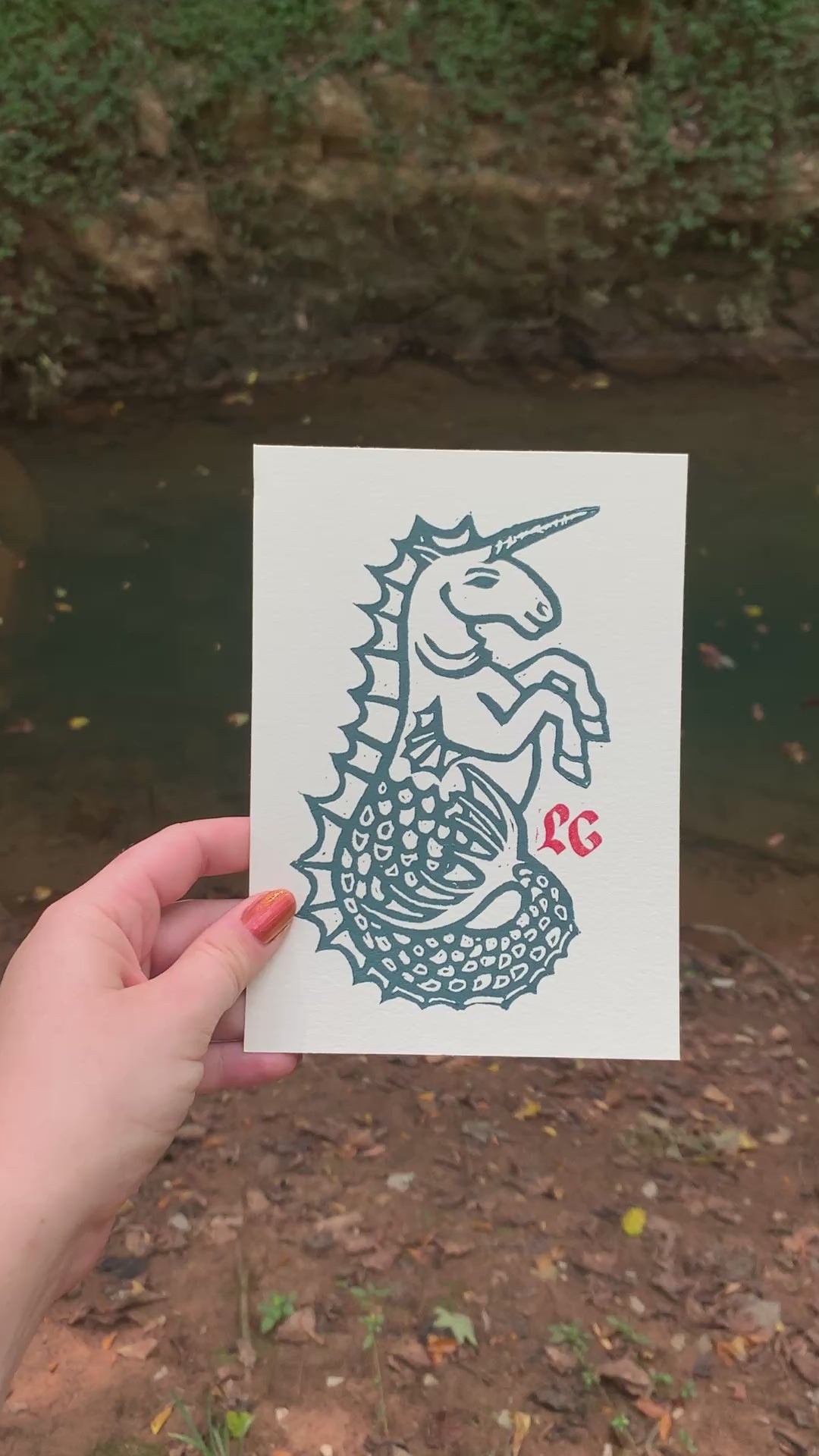 A hand with orange nail polish holding the Hippocampus Art Print in front of a Kentucky blue hole. The blue ink of the art print and the blue of the blue hole matches.